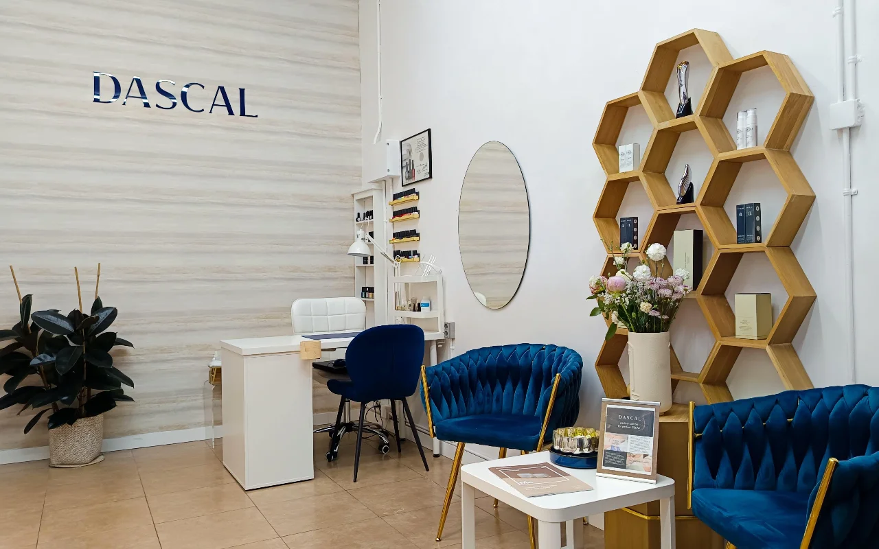 Un intérieur de salon de beauté élégant avec des fauteuils en velours bleu nuit et une étagère murale hexagonale en bois. Un bureau de manucure blanc est installé dans un coin contre un mur texturé clair, avec le mot  DASCAL  en lettres bleues.