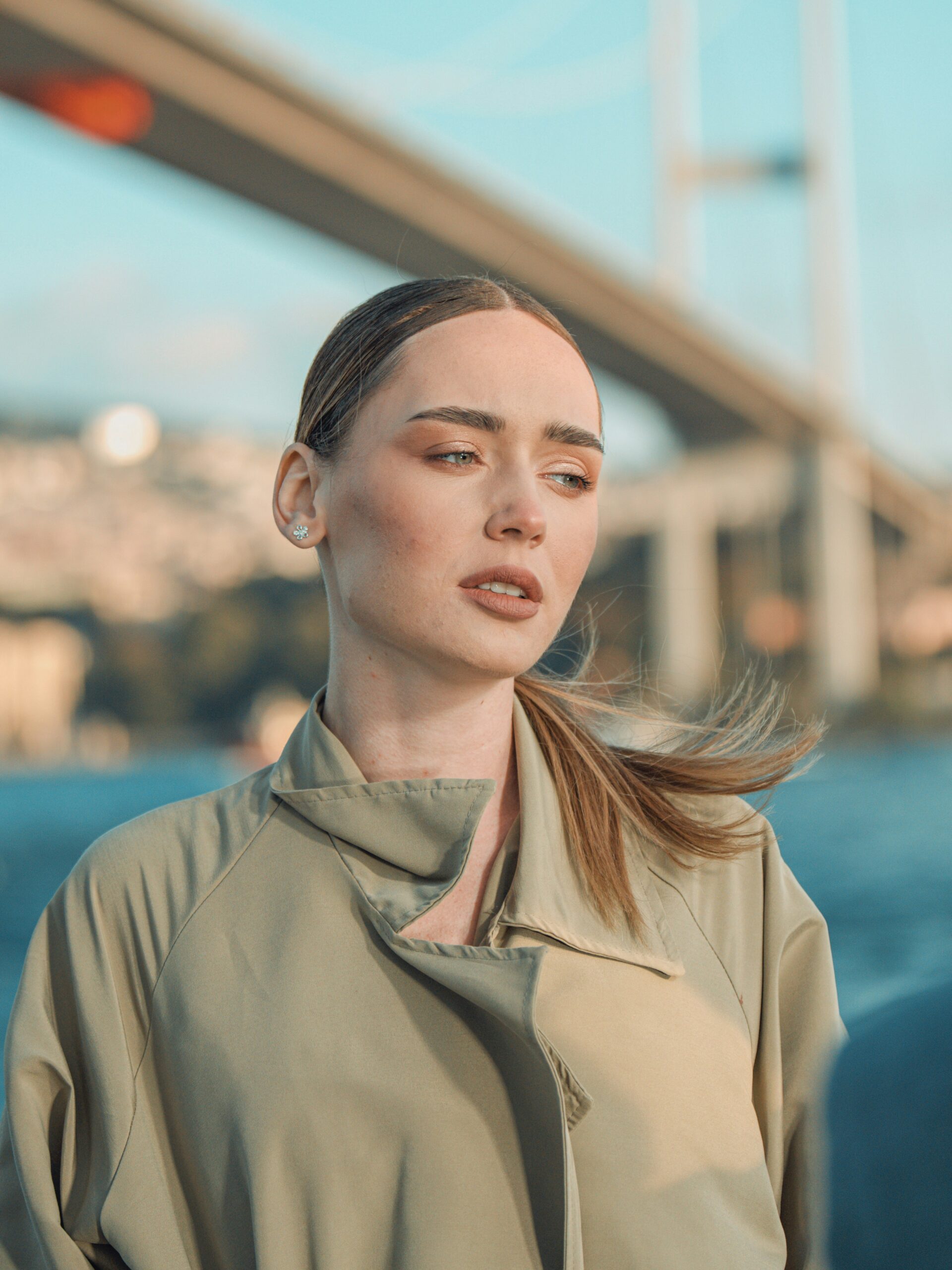 Une femme avec un microblading des sourcils et des cheveux clairs attachés, portant un trench, debout devant un pont à Istanbul