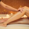 Vue latérale des mains d’une esthéticienne lissant délicatement les jambes d’une femme allongée sur une table de soins dans un salon après une épilation à la cire