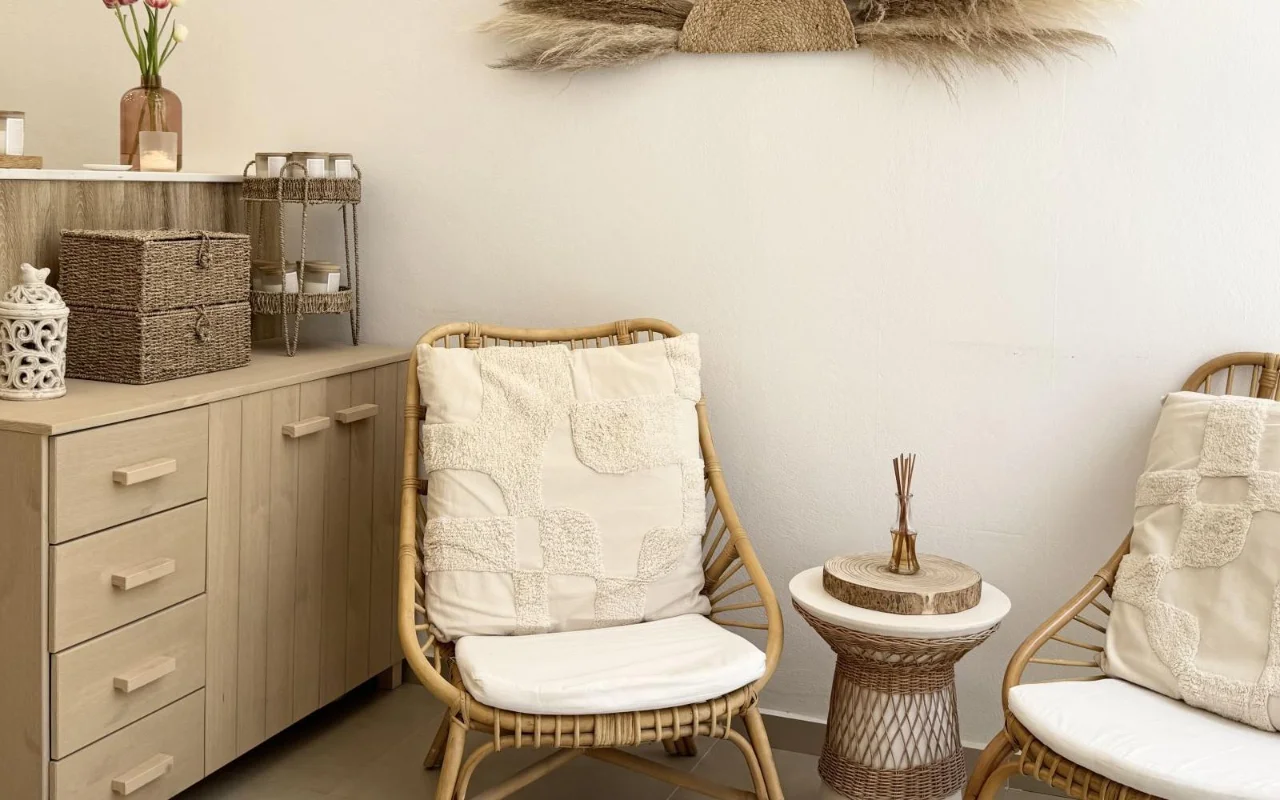 Un espace d’attente apaisant avec des fauteuils en rotin, des coussins crème texturés, une table d’appoint en osier avec un diffuseur, des paniers de rangement tressés, et de l’herbe de la pampa séchée au mur au-dessus d’un meuble en bois clair.