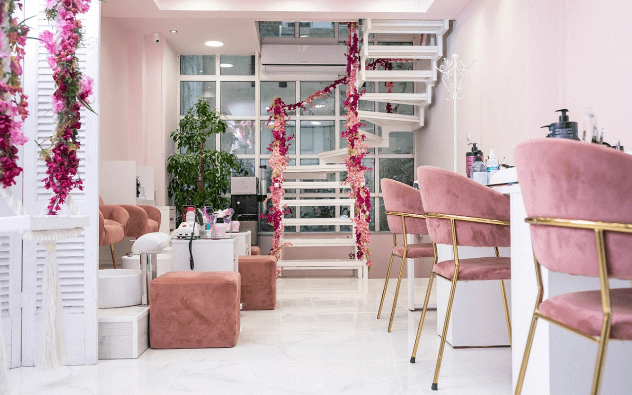 Un salon de manucure féminin avec des fauteuils en velours rose poudré, un escalier en colimaçon blanc orné de fleurs artificielles roses, des cloisons à persiennes, et des postes de manucure aux pieds dorés sur un sol effet marbre.