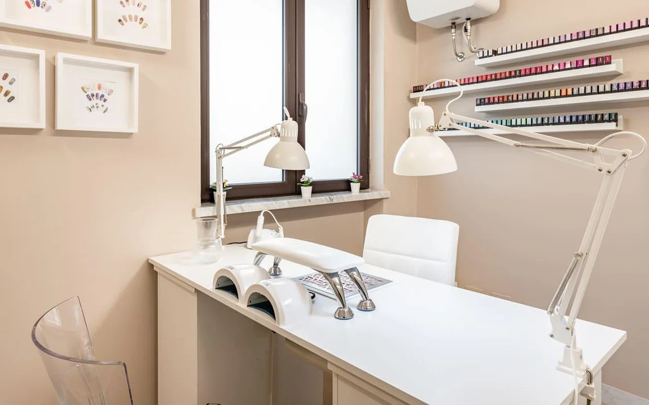Un poste de manucure dédié chez AG The Beauty Room's, avec un bureau blanc épuré, deux lampes UV, des liseuses réglables et des étagères présentant des rangées de vernis colorés. Des créations de nail art encadrées ornent les murs aux tons chaleureux.