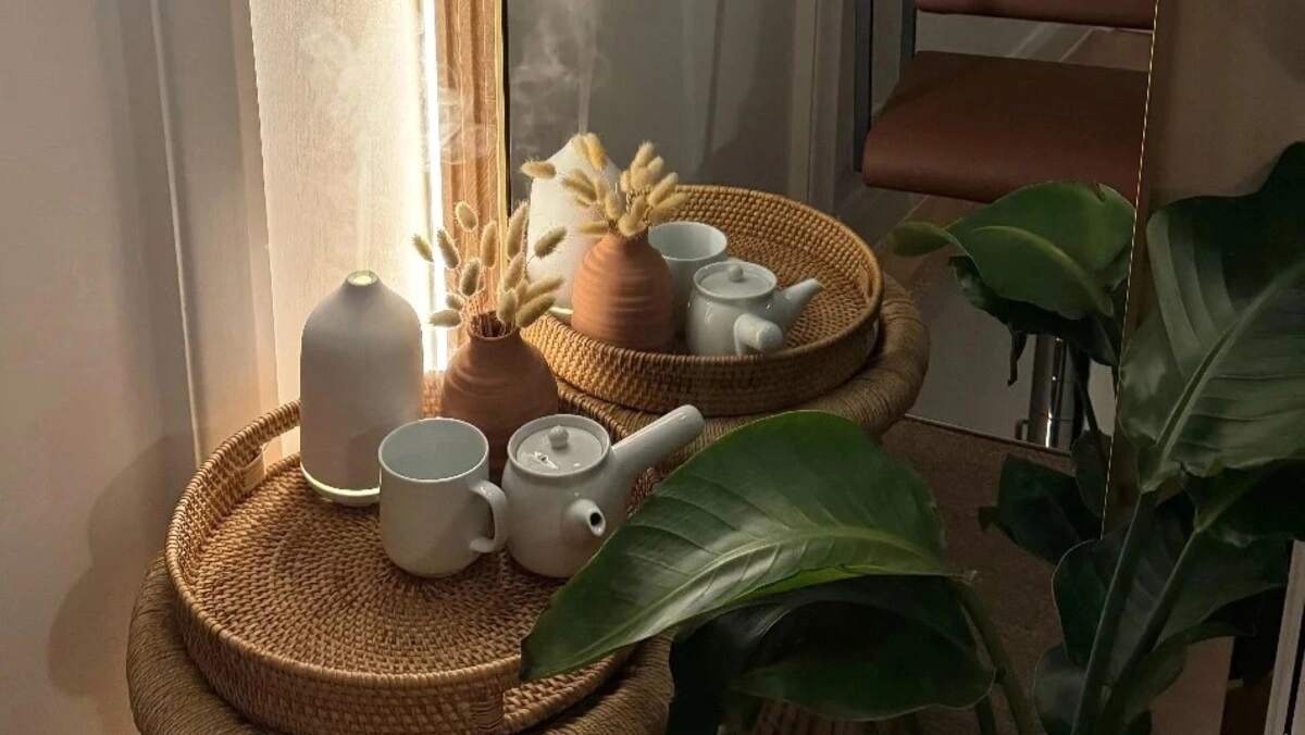 Gros plan d'un plateau en osier avec un diffuseur et un service à thé à côté d'une grande plante verte dans un salon