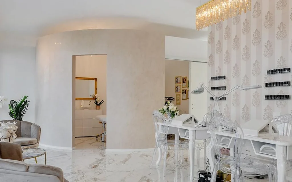 L'intérieur élégant du MC Beauty Center, avec un sol effet marbre, des murs blancs incurvés et un lustre en cristal doré. Un poste de manucure avec des chaises transparentes se trouve à droite, tandis que des fauteuils en velours et une porte au cadre doré mènent à une cabine de soin à l'arrière.