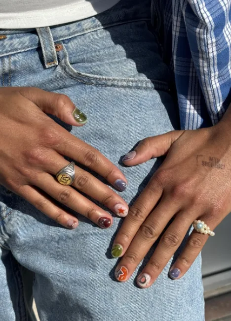 Manucure dépareillée sur ongles courts, chaque ongle d'une couleur et d'un motif différents, avec une étoile rouge et une volute orange, posée sur un jean.