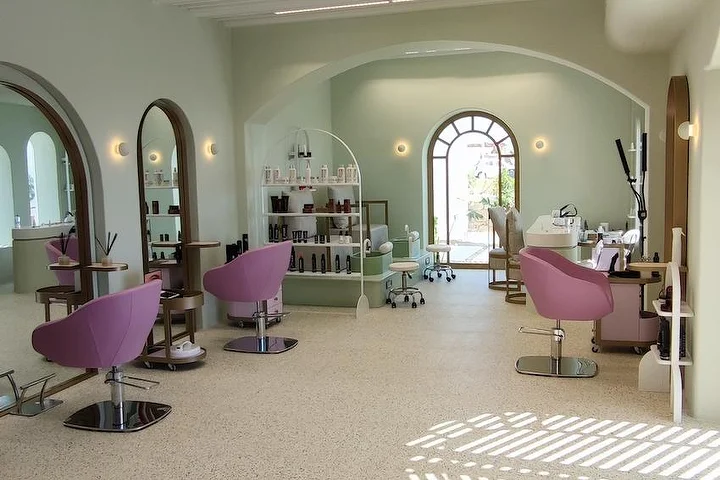 Un salon lumineux vert sauge au sol en terrazzo, avec des miroirs cintrés à cadre de bois et une fenêtre en arc donnant sur le jardin, des fauteuils de coiffage rose poudré et des étagères ouvertes garnies de produits, la lumière du soleil tombant sur le sol.