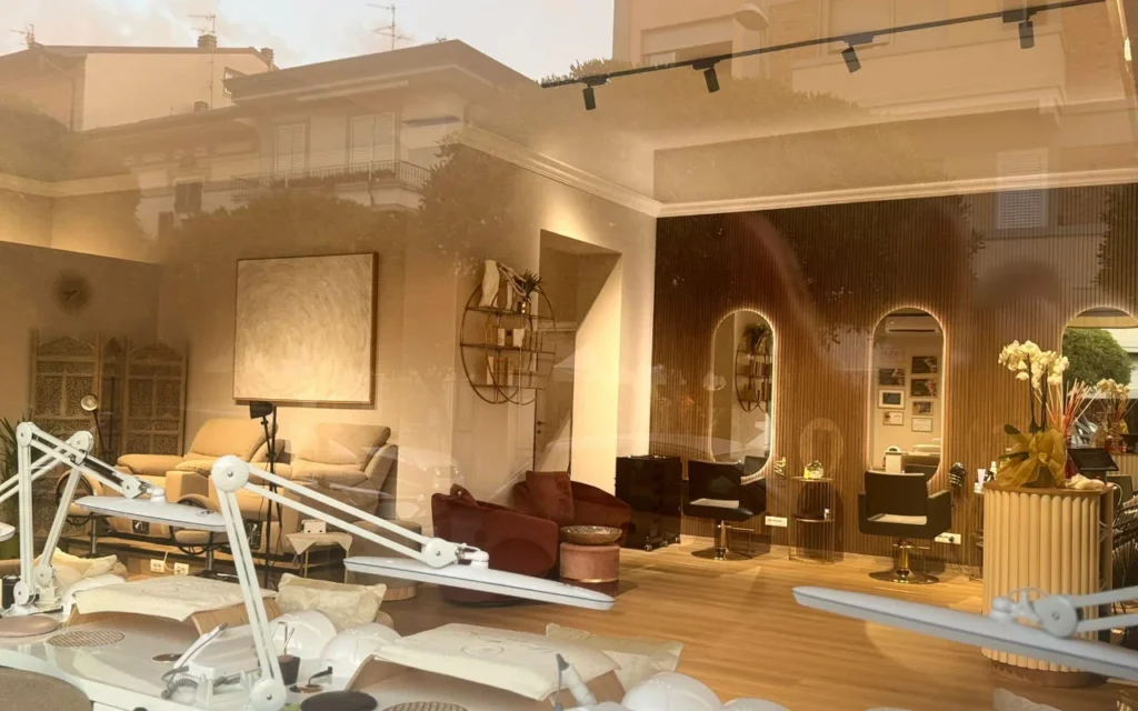 L'intérieur du salon de Lilian Almanza vu à travers la vitrine à l'heure dorée. Des postes de manucure équipés de lampes réglables s'alignent à gauche, tandis que la droite révèle des boiseries chaleureuses, des miroirs cintrés aux cadres rétroéclairés et des fauteuils en velours.