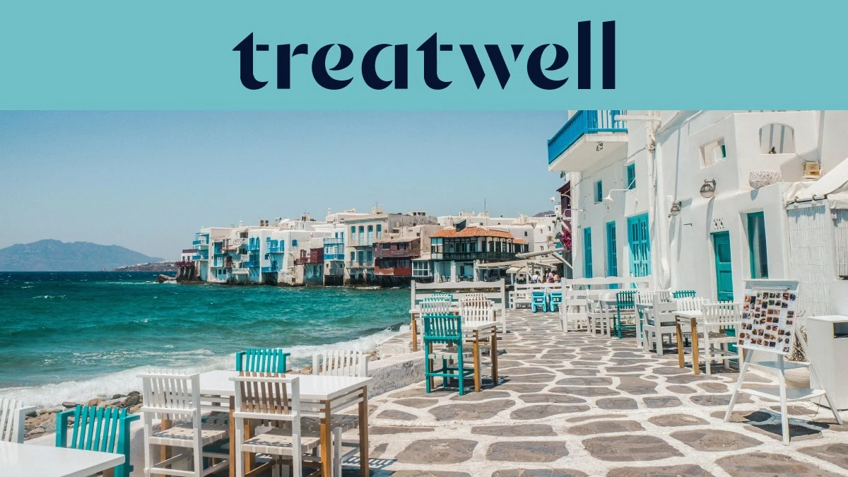 Le front de mer de la Petite Venise à Mykonos : maisons blanchies à la chaux aux balcons de bois colorés au bord de l'eau, la mer Égée turquoise au loin, et au premier plan des tables de café blanches et bleu canard installées sur une terrasse pavée.