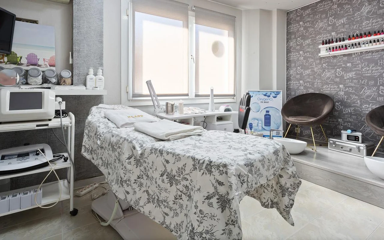 Une cabine de soin lumineuse avec une table habillée d'un linge à motifs floraux gris et de serviettes blanches pliées, des appareils d'esthétique sur chariots à gauche, une fenêtre au store à moitié baissé, et un espace surélevé avec un fauteuil en velours marron et des bacs de pédicure devant un mur d'accent à motif manuscrit.