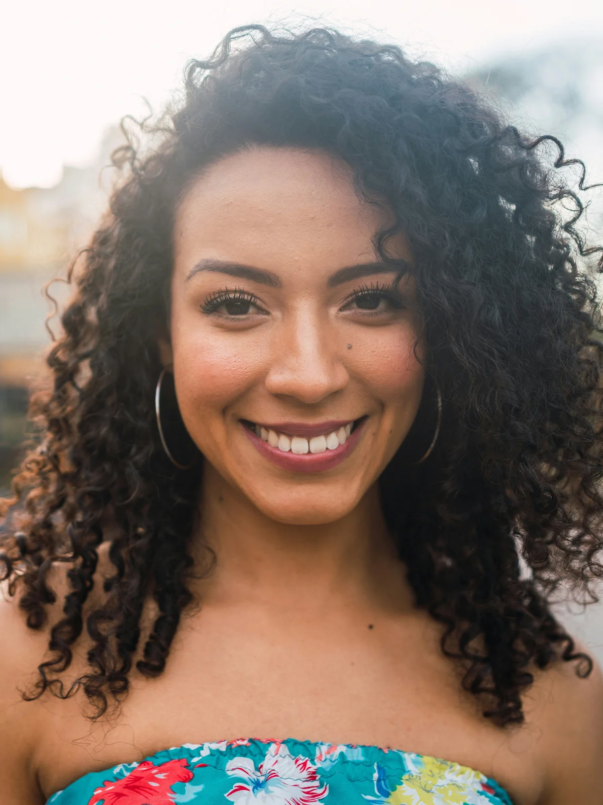 Une femme aux sourcils parfaitement dessinés sourit face à l'objectif, vêtue d'un top bustier à fleurs, dehors dans la lumière du soir