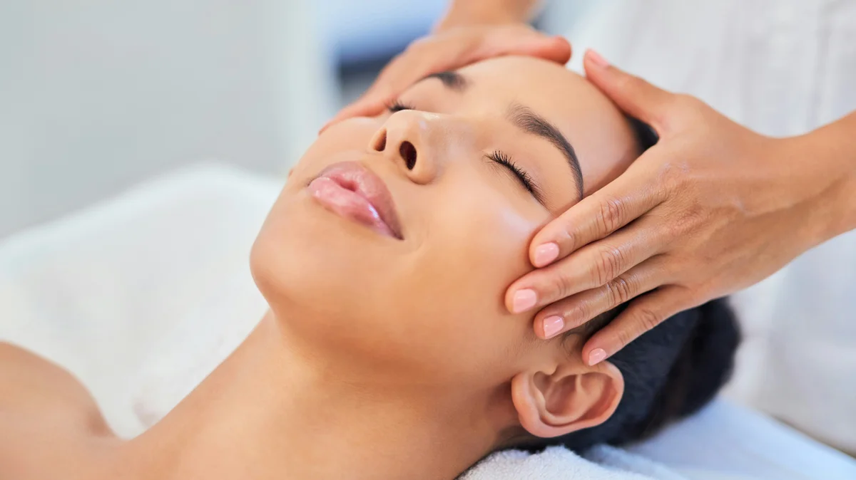 Une femme allongée dans un institut de beauté pendant qu'une esthéticienne pose délicatement les mains sur sa tête lors d'un soin du visage