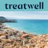 Vue panoramique sur une petite ville côtière sicilienne, avec de vieux bâtiments aux toits de terre cuite, un croissant de plage de sable, une mer turquoise limpide et un promontoire rocheux qui s'élève sur un côté.