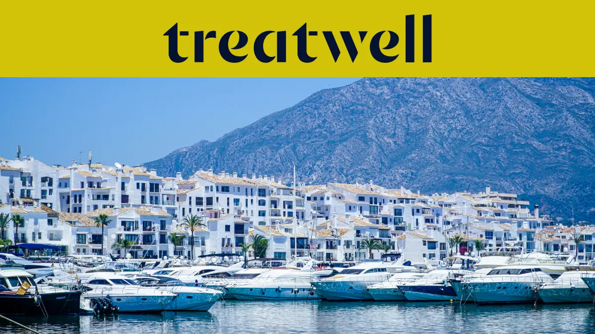 Le port de plaisance de Puerto Banús, à Marbella, bordé d'immeubles blancs et de yachts amarrés sur fond de montagnes, sous une banderole jaune Treatwell.