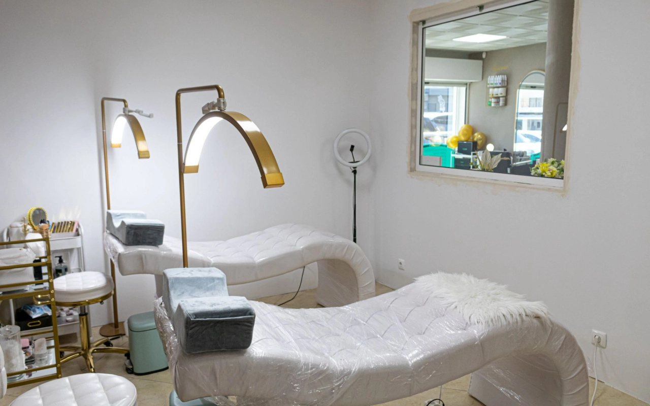 Une salle de soins beauté lumineuse avec deux transats blancs matelassés, de hauts lampadaires arqués dorés et une lampe annulaire, avec une fenêtre intérieure donnant sur la réception.