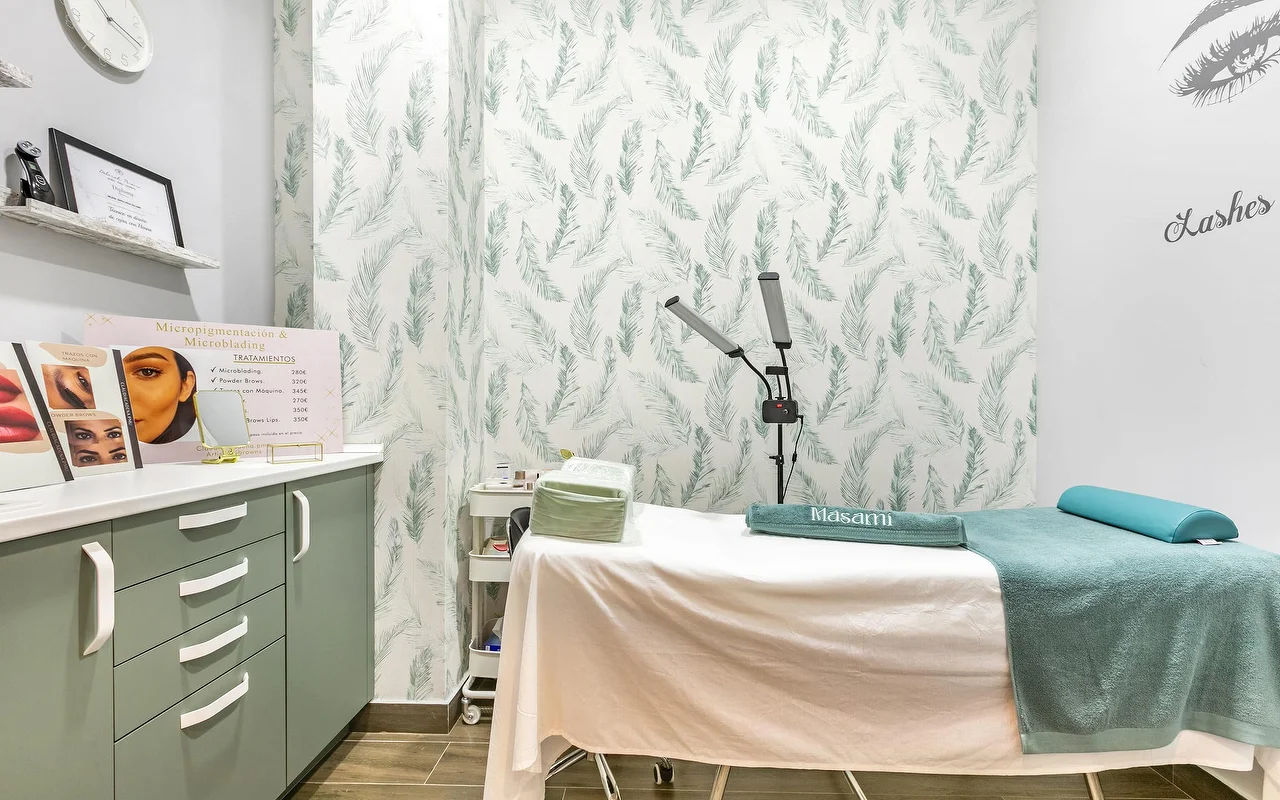 Une cabine de soin chez Masami Belleza Natural à Fuengirola, avec une table de soin, un papier peint à motif de plumes, une lampe-loupe et un affichage de tarifs de micropigmentation.