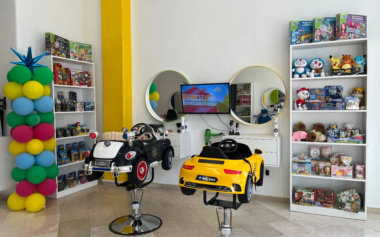 L'espace enfants de Peluplay à Benalmádena, où les fauteuils de coiffage ont la forme de petites voitures, avec des étagères pleines de dessins animés, des miroirs ronds et une colonne de ballons.