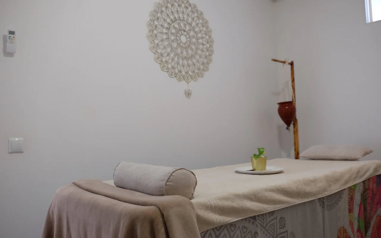 Une salle de massage apaisante dans des tons neutres et doux, avec une table de massage drapée d'une serviette et un traversin, un grand mandala au crochet accroché au mur, et un support en bois portant un pot en terre cuite suspendu.