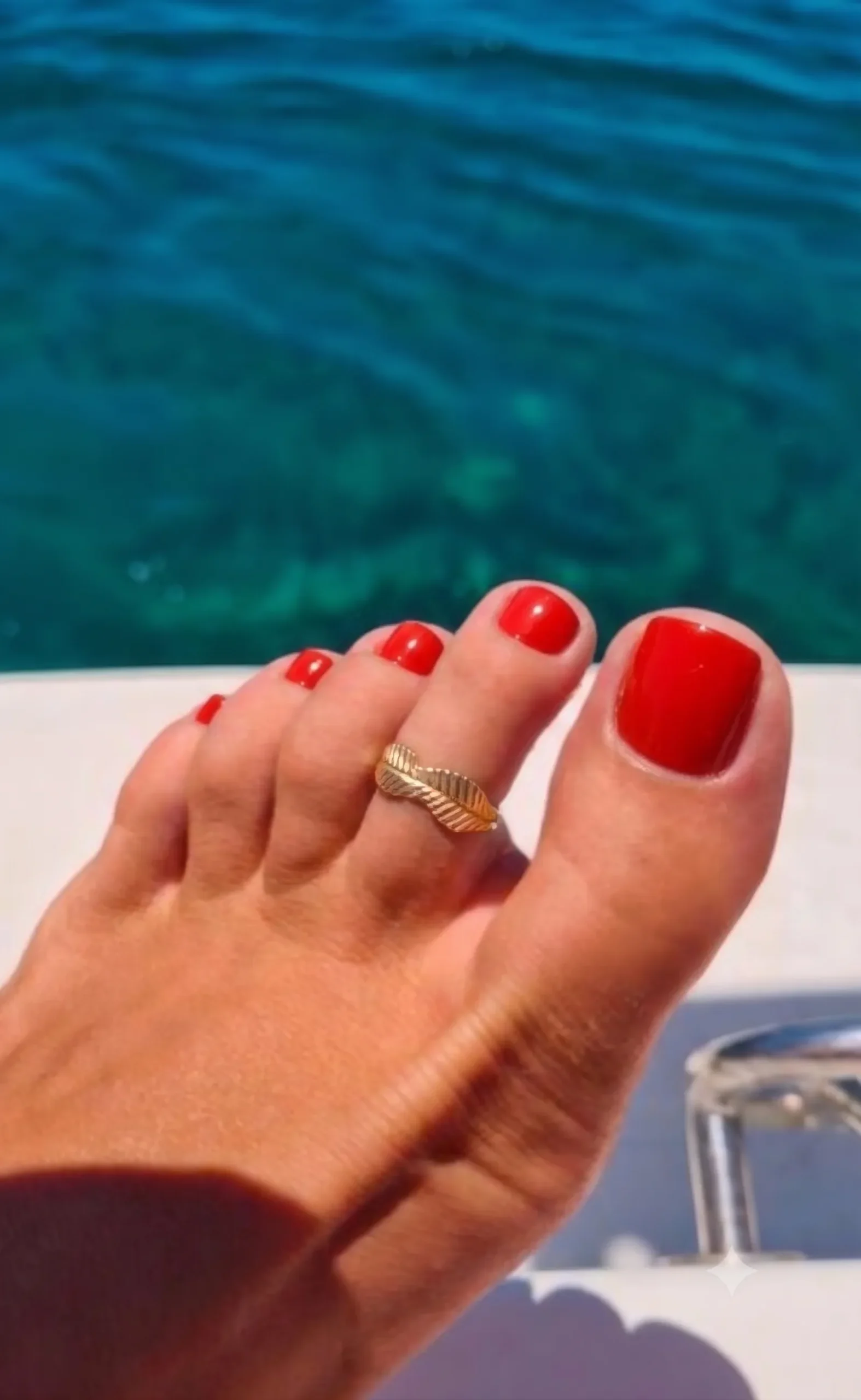 Pédicure rouge tomate brillante sur un pied hâlé avec une bague d'orteil dorée, mer turquoise en arrière-plan