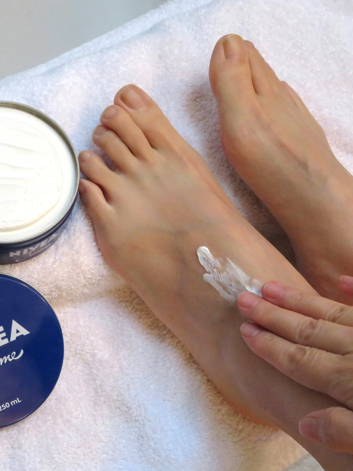 Gros plan de dessus sur les pieds d'une femme posés sur un drap blanc, un pot de crème ouvert à côté, pendant qu'elle applique sa crème