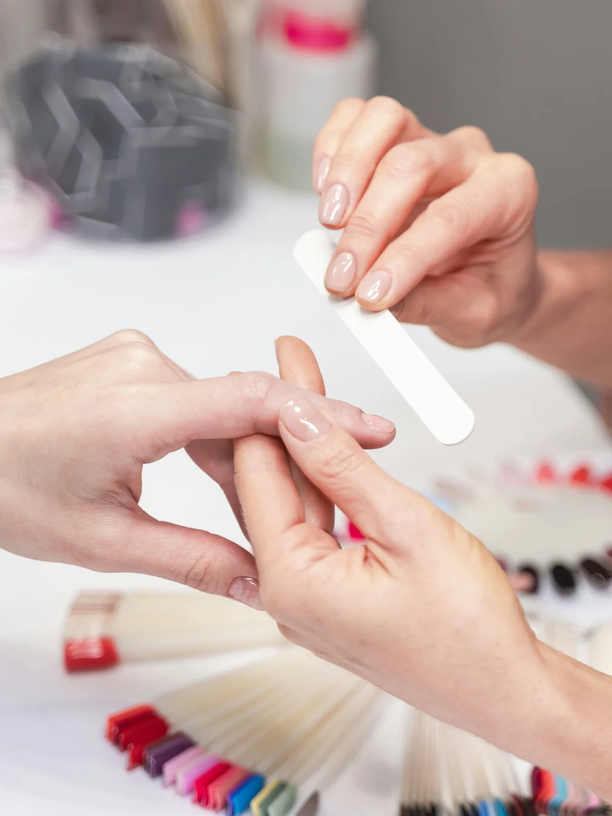 Gros plan sur une prothésiste qui lime doucement les ongles d'une cliente dans un salon, avec du matériel et des produits de manucure en arrière-plan