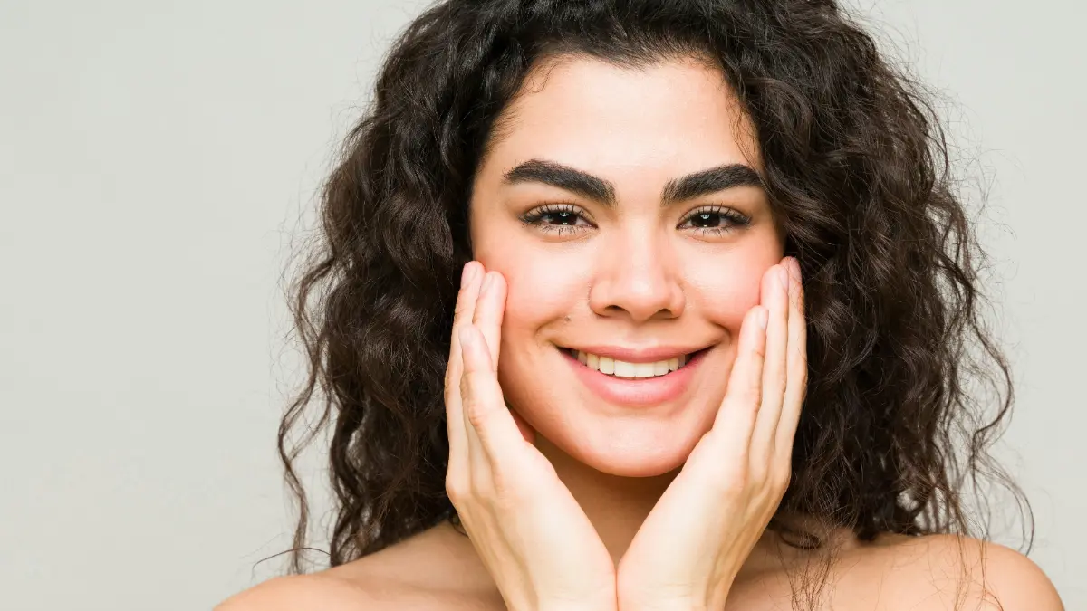 Une jeune femme à la peau nette et aux cheveux foncés ondulés sourit face à la caméra