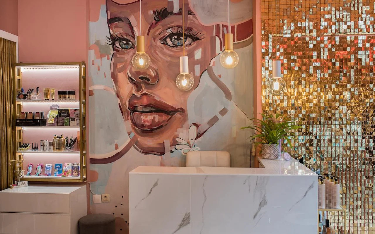 Un salon aux murs roses avec une grande fresque peinte représentant un visage de femme, un mur de paillettes dorées scintillantes, un comptoir d'accueil en marbre blanc et des suspensions à ampoules apparentes.