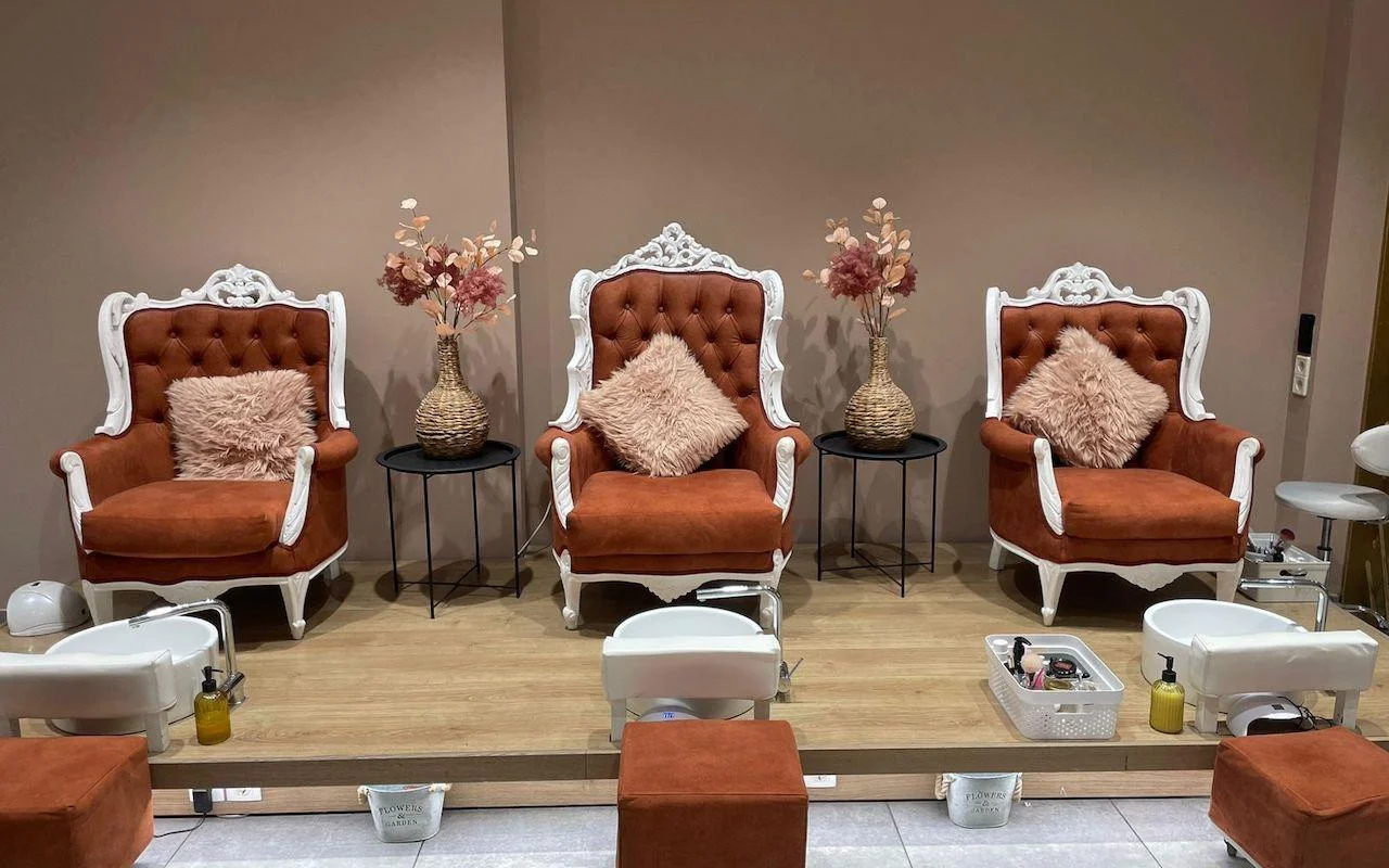 Trois fauteuils ornés à cadre blanc en velours orange brûlé, surélevés sur une plateforme en bois au-dessus de bassines pédicure blanches, avec des fleurs séchées et des coussins rose tendre disposés entre les sièges.