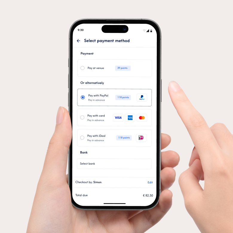 Primo piano di uno smartphone che mostra l'interfaccia di pagamento dell'app Treatwell per la selezione del metodo di pagamento. Tra le opzioni ci sono il pagamento in salone o il pagamento anticipato con PayPal, carta di credito o iDeal. Una mano tiene il telefono e un dito ne indica lo schermo.