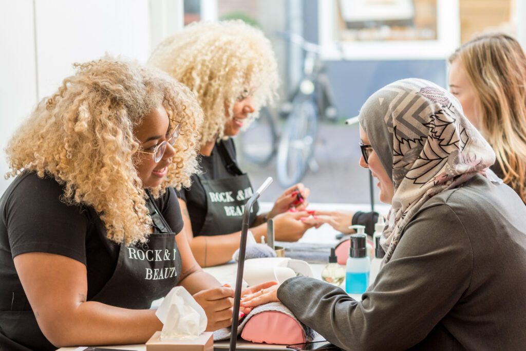 Due manicuriste con capelli biondi ricci e grembiuli neri con la scritta "Rock & Roll Beauty" fanno la manicure a delle clienti su un lungo tavolo. Una cliente indossa un hijab grigio.