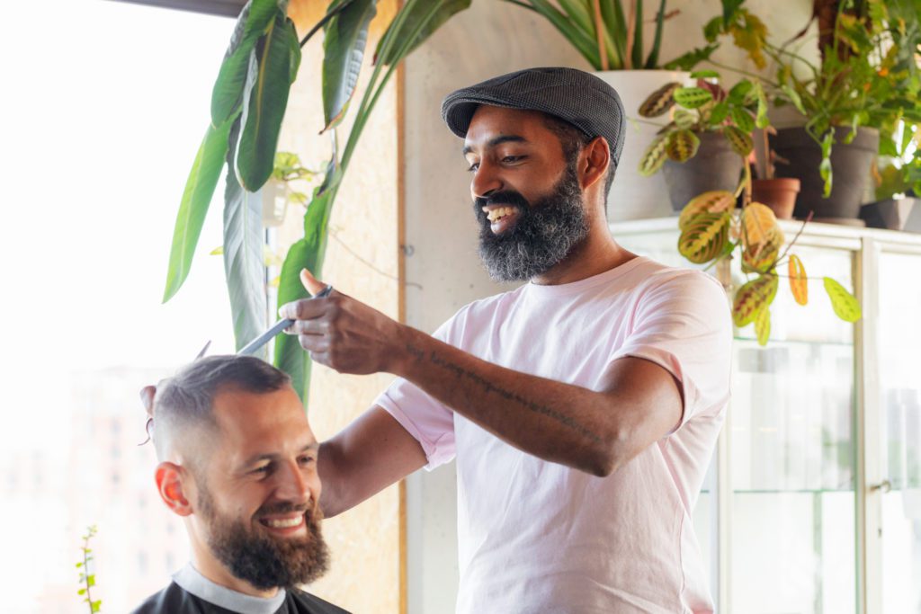 Un barbiere sorridente con la barba scura e una coppola grigia usa forbici e pettine sui capelli di un cliente sorridente in un salone luminoso ricco di piante verdi da interno.