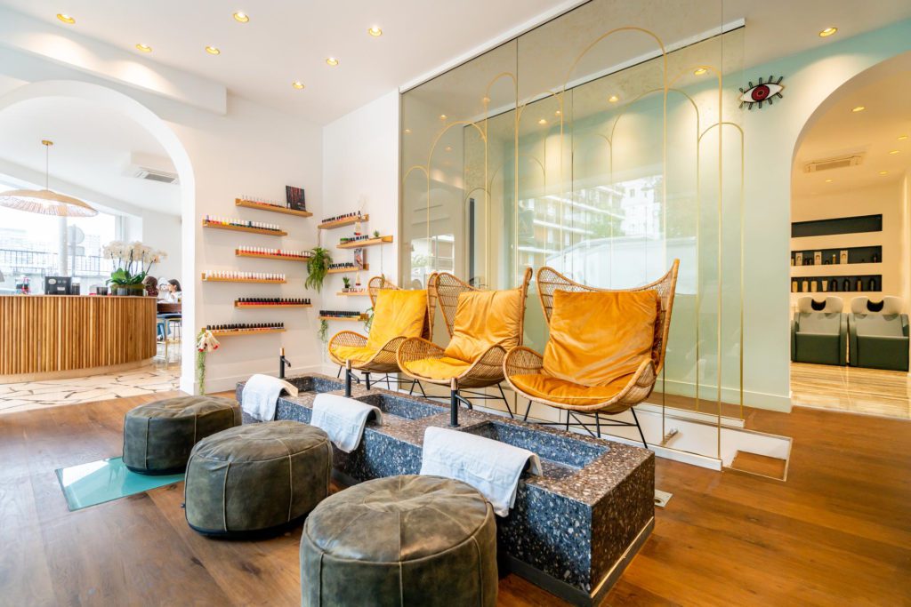 Interno elegante e luminoso di un salone di bellezza che mostra una postazione pedicure con tre poltrone in vimini color giallo senape, vaschette in pietra con asciugamani bianchi e pouf grigi coordinati su un pavimento in legno.