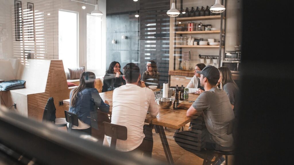 Vista spontanea attraverso una vetrata di un gruppo di colleghi seduti attorno a un lungo tavolo in legno nella cucina o zona pranzo di un ufficio moderno, mentre chiacchierano e bevono qualcosa insieme.