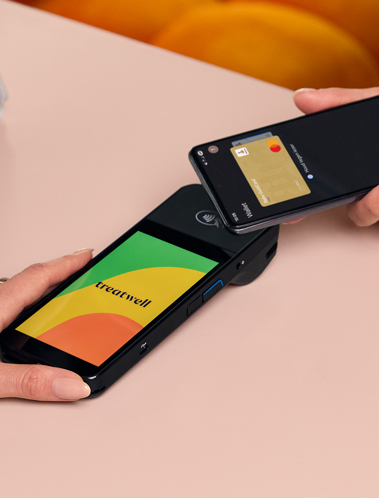 Una mano tiene un terminale di pagamento Treatwell nero mentre un cliente avvicina lo smartphone con wallet digitale per pagare in modalità contactless.