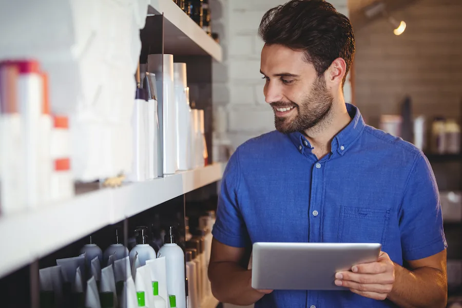 Un titolare di salone sorridente in camicia blu guarda un tablet accanto a scaffali di prodotti di bellezza.