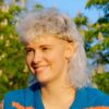 Una giovane donna con un mullet riccio biondo platino, seduta all'aperto in una giornata di sole.