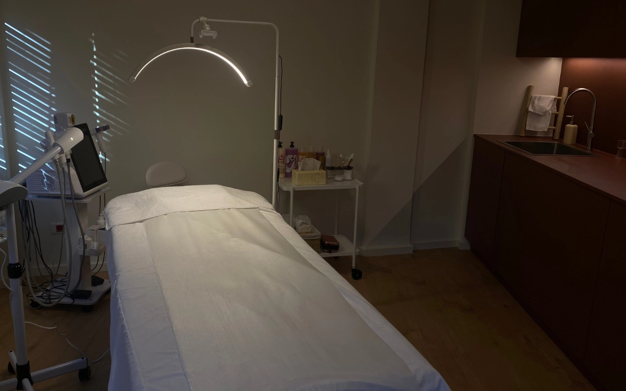 Una sala trattamenti essenziale con un lettino bianco sotto una lampada ad arco a LED, apparecchiature estetiche professionali con schermo digitale, prodotti per la cura della pelle su un carrello e un mobile in legno scuro con lavandino lungo la parete di fondo.