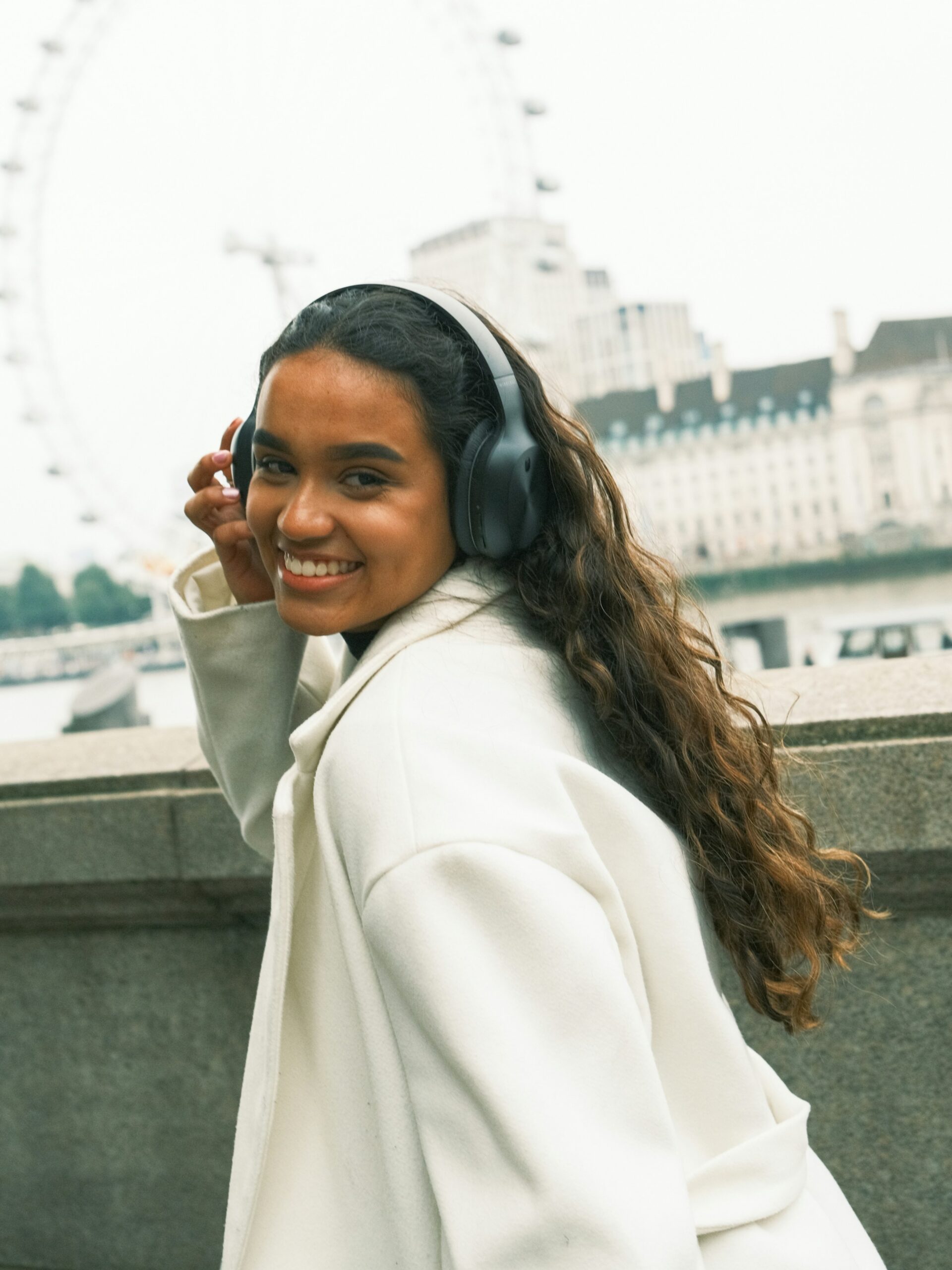 Una donna sorridente con microblading sulle sopracciglia, cuffie nere over-ear e cappotto bianco, in una città svizzera
