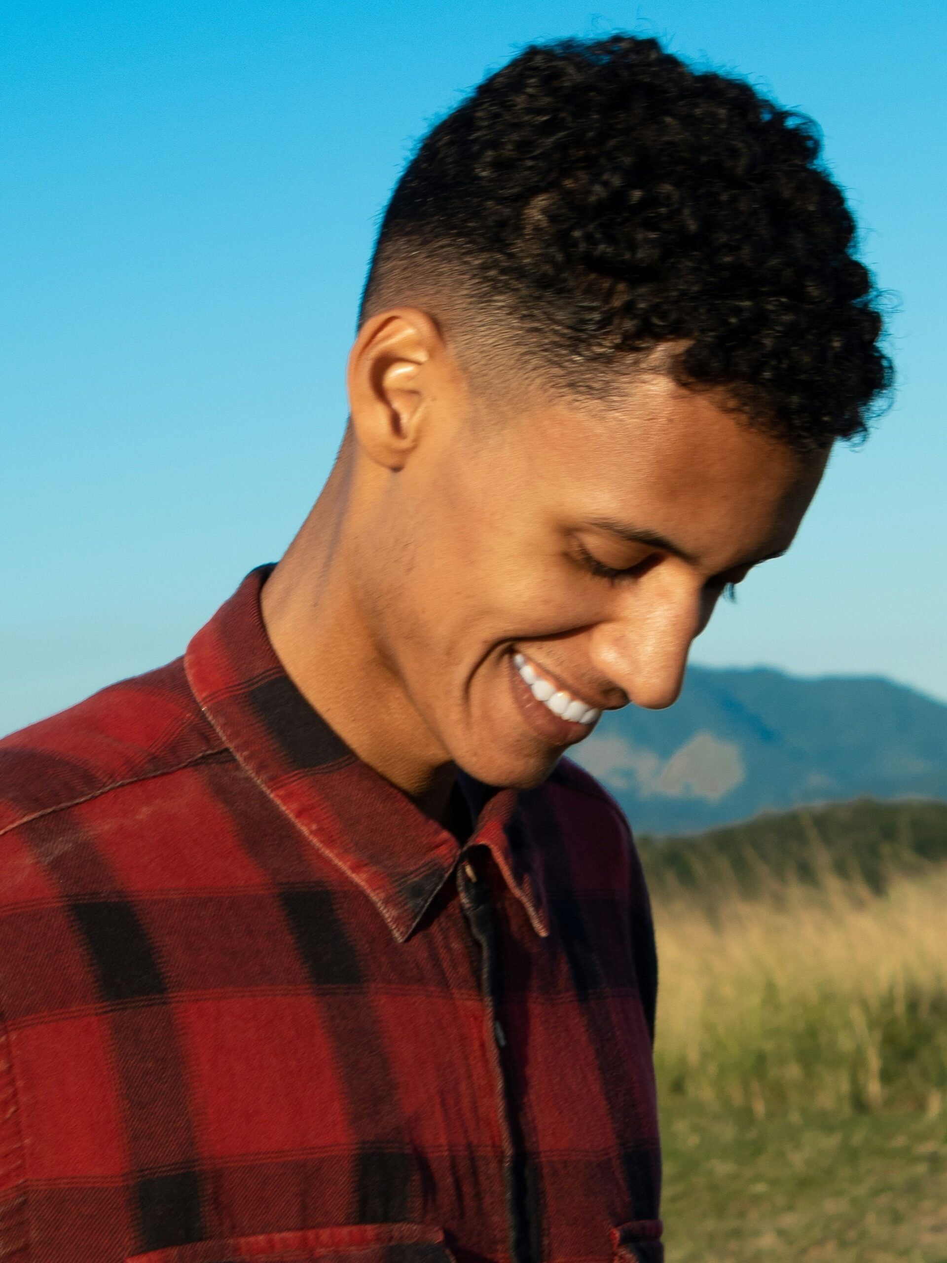 Uomo sorridente all'aperto con sfondo di montagne e cielo azzurro, con capelli ricci e un taper fade medio