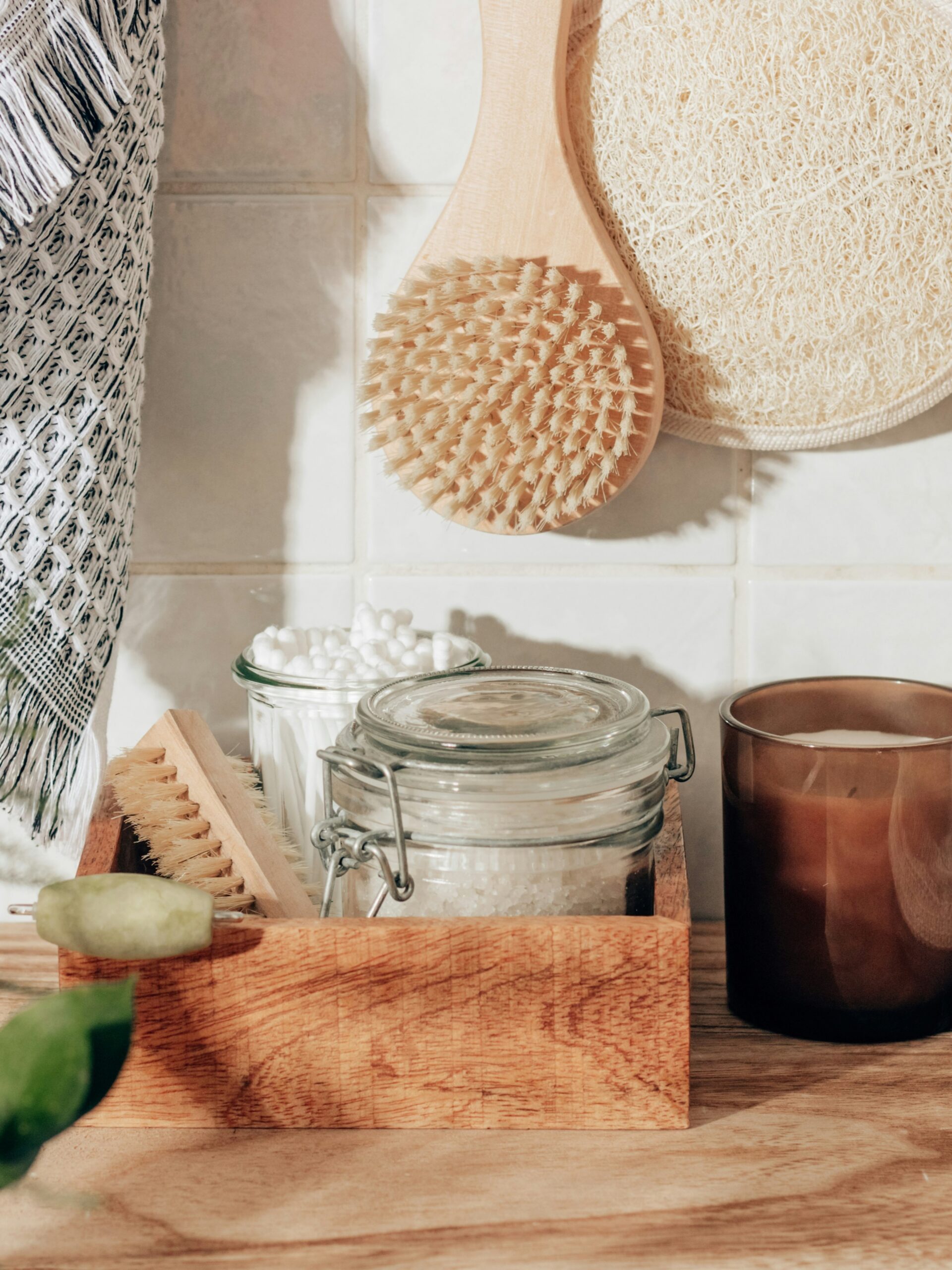 Accessori da bagno disposti con cura: guanto esfoliante, spazzola corpo, sali da bagno, dischetti di cotone e una candela