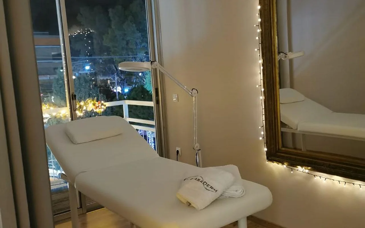 Una sala trattamenti con luce soffusa, lettino bianco, asciugamano con il marchio CasaDerm, una lampada ingranditrice, lucine decorative intorno a uno specchio dorato e una porta-finestra con vista sulla via serale di Glyfada.