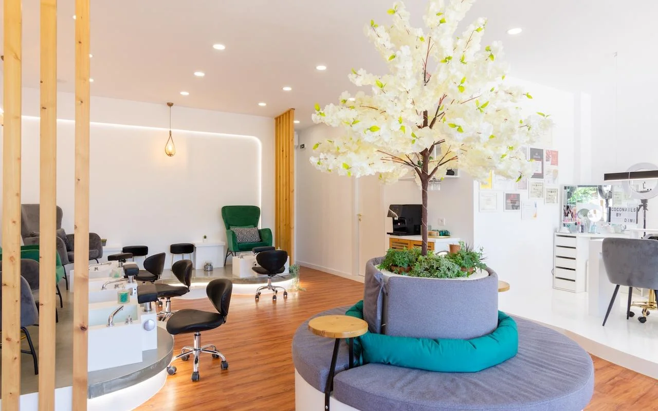 Un salone unghie aperto e dal tocco naturale con un grande albero decorativo bianco in fiore come punto focale, poltrone in velluto verde smeraldo alle postazioni pedicure, divisori a listelli di legno e un pavimento in legno caldo.