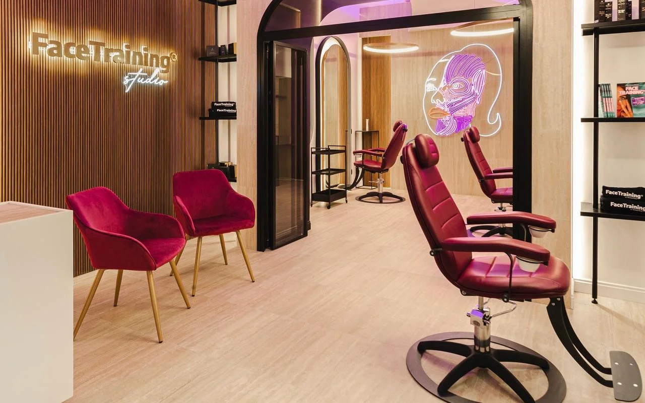 Uno studio minimalista con una parete a doghe di legno e un logo retroilluminato. L'area trattamenti comprende poltrone professionali in pelle bordeaux e un'installazione luminosa al neon che raffigura un diagramma stilizzato dei muscoli facciali su una parete divisoria in vetro.
