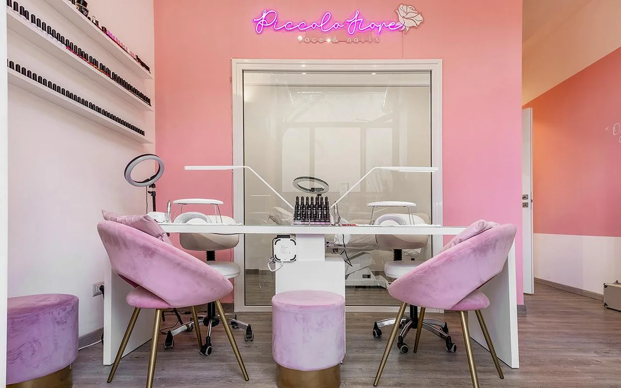 Una postazione manicure con pareti rosa acceso e un'insegna al neon fucsia. Sedie in velluto lilla con gambe dorate sono posizionate davanti a una scrivania bianca e minimalista con lampade LED professionali per unghie.