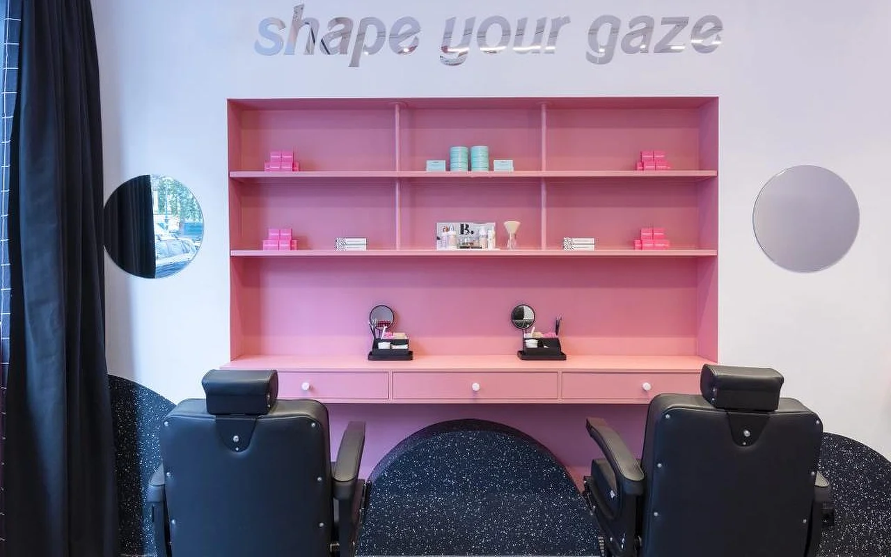 Uno studio specializzato in sopracciglia e ciglia con una vistosa scaffalatura rosa che espone prodotti beauty, la scritta argentata 'shape your gaze' sulla parete, poltrone nere in pelle per i trattamenti e un pavimento scuro a effetto granito.