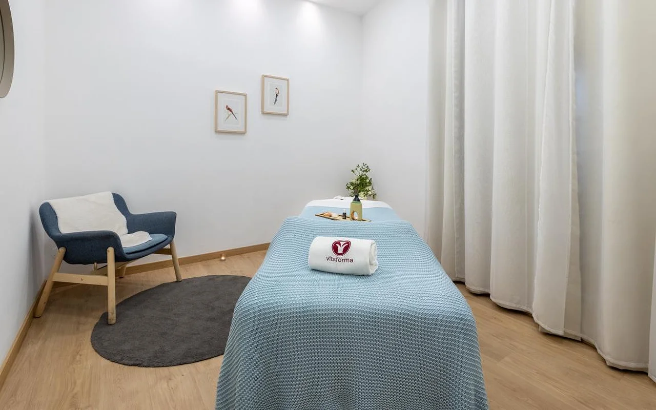 Una stanza spa minimalista e pulita con pareti bianche e pavimento in legno chiaro. Un lettino da trattamento è ordinatamente preparato con una coperta a maglia azzurra e un asciugamano bianco arrotolato con il logo 'Vitaforma'. In un angolo, un'elegante poltrona blu poggia su un piccolo tappeto rotondo grigio, e lunghe tende bianche incorniciano la finestra.