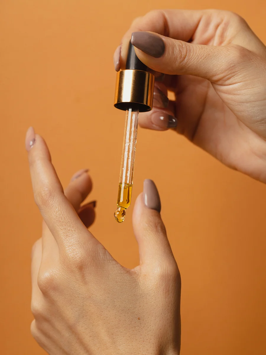 Primo piano di due mani con manicure, una delle quali tiene un contagocce di vetro con l’olio per cuticole che gocciola