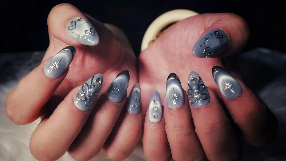 Primo piano delle mani di una donna con unghie acriliche elaborate dall’effetto occhio di gatto grigio, cuori e stelle argentati e decori con strass scintillanti