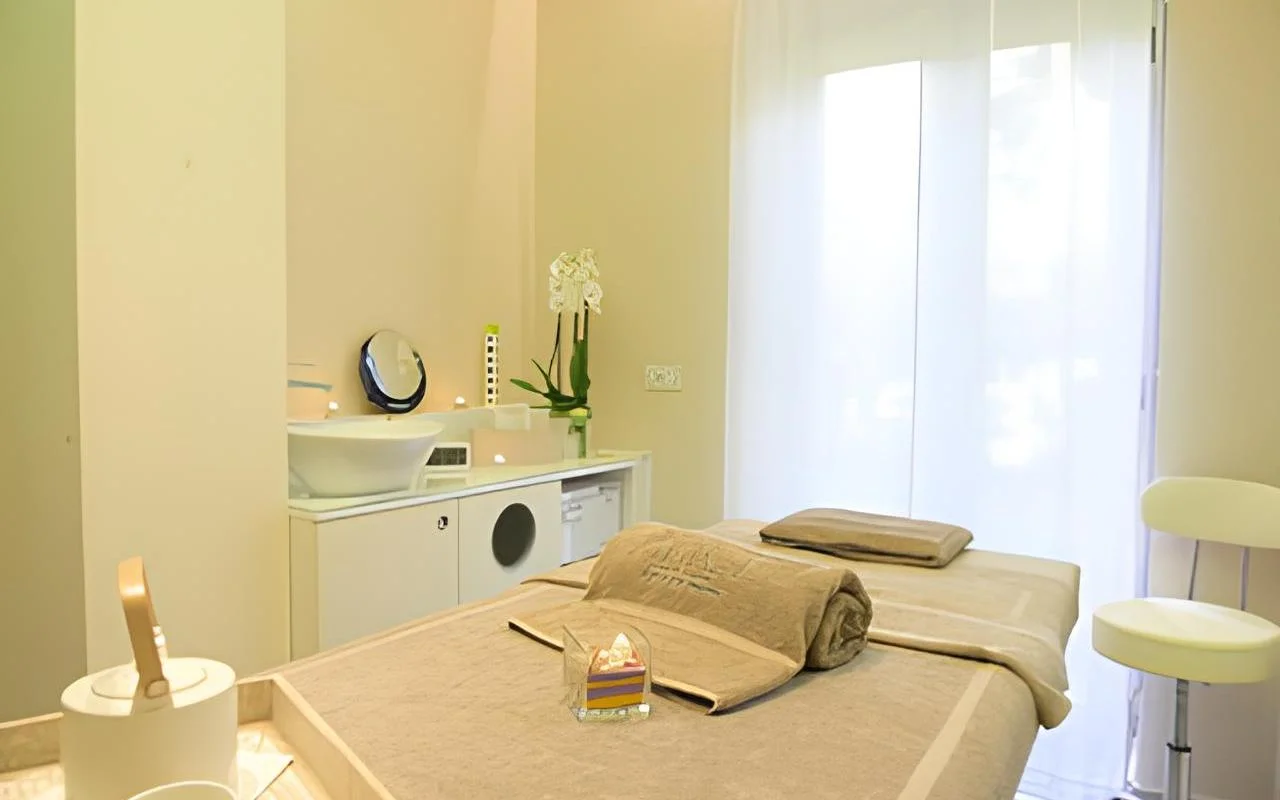 Una cabina trattamenti rilassante da Alice Beauty Clinique, con un lettino vestito di lino color tortora tenue, orchidee bianche sul piano, candele accanto al lavabo e tende leggere che filtrano la luce naturale da una grande finestra.