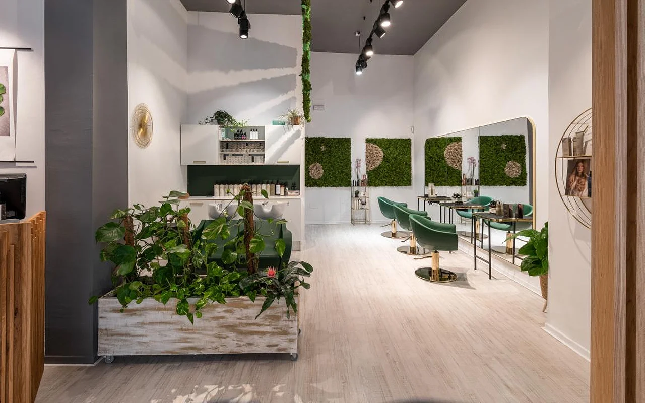 Salone di parrucchiere pieno di piante, con pavimenti in legno chiaro, una fioriera bianca anticata traboccante di verde, un quadro di muschio verde alla parete, poltrone da styling in velluto verde smeraldo con dettagli dorati e una postazione di lavaggio sul fondo.