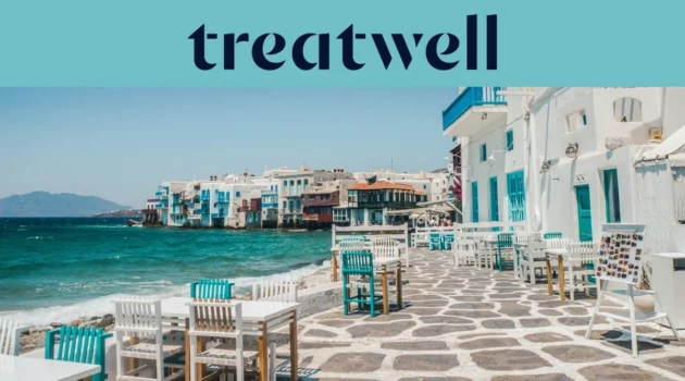 Il lungomare della Piccola Venezia a Mykonos: case imbiancate a calce con balconi di legno colorati a filo dell'acqua, il mar Egeo turchese sullo sfondo e, in primo piano, tavolini da caffè bianchi e verde acqua su una terrazza lastricata in pietra.